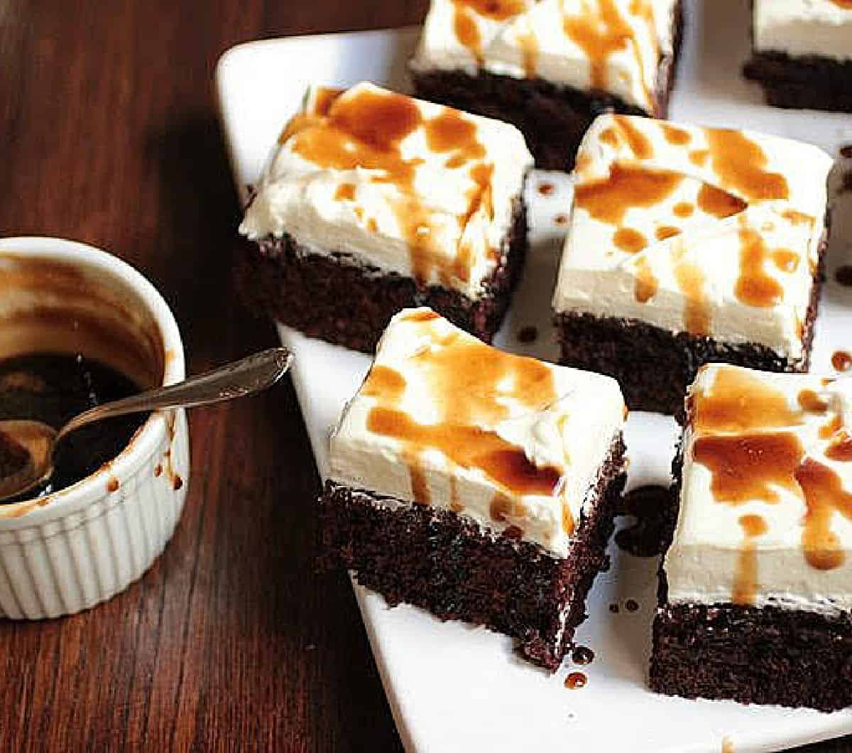 Wooden table with chocolate cake squares topped with whipped cream and syrup; a white ramekin beside