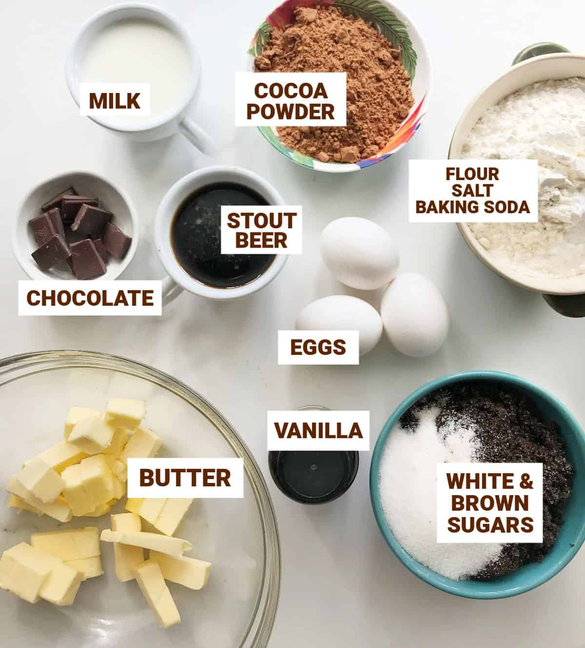 Different bowls with ingredients for chocolate stout cake on a white surface, including cocoa, eggs and butter