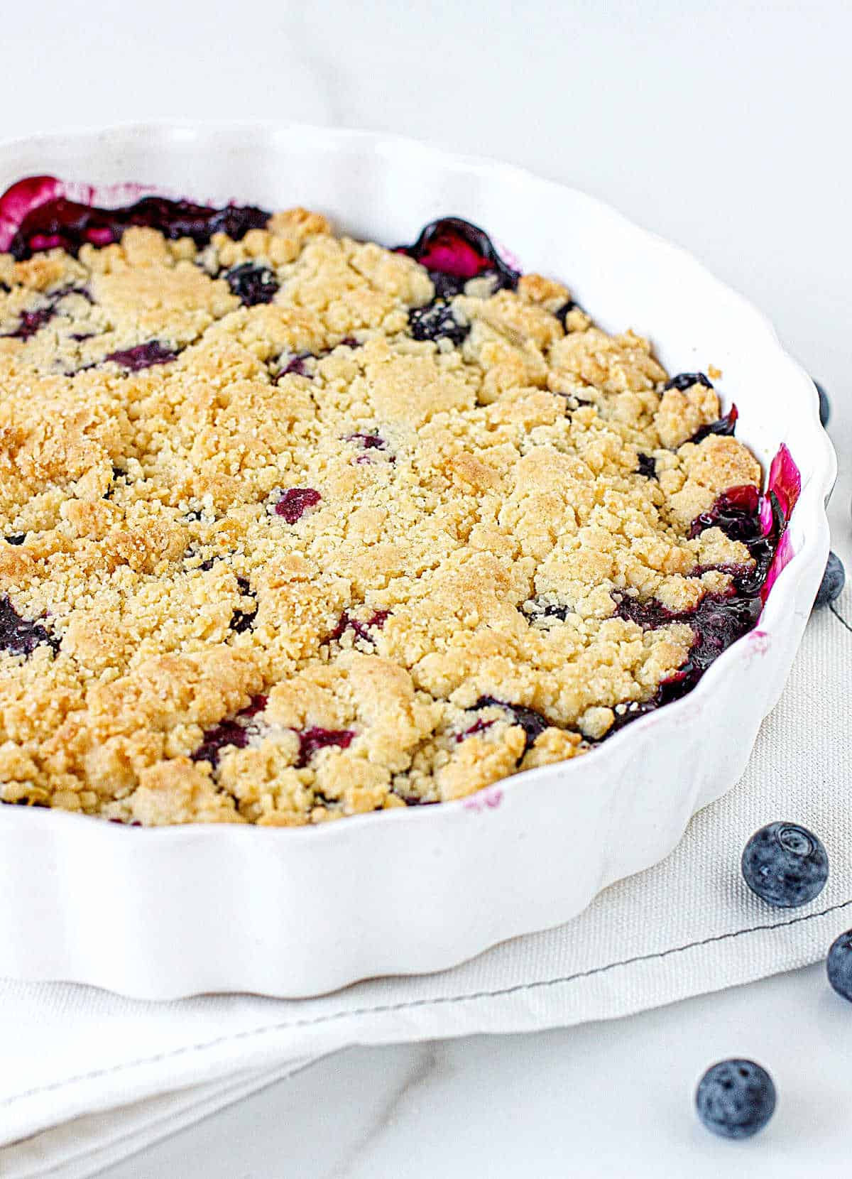 Round white baking dish with baked blueberry dump cake on a white cloth with a white background. 