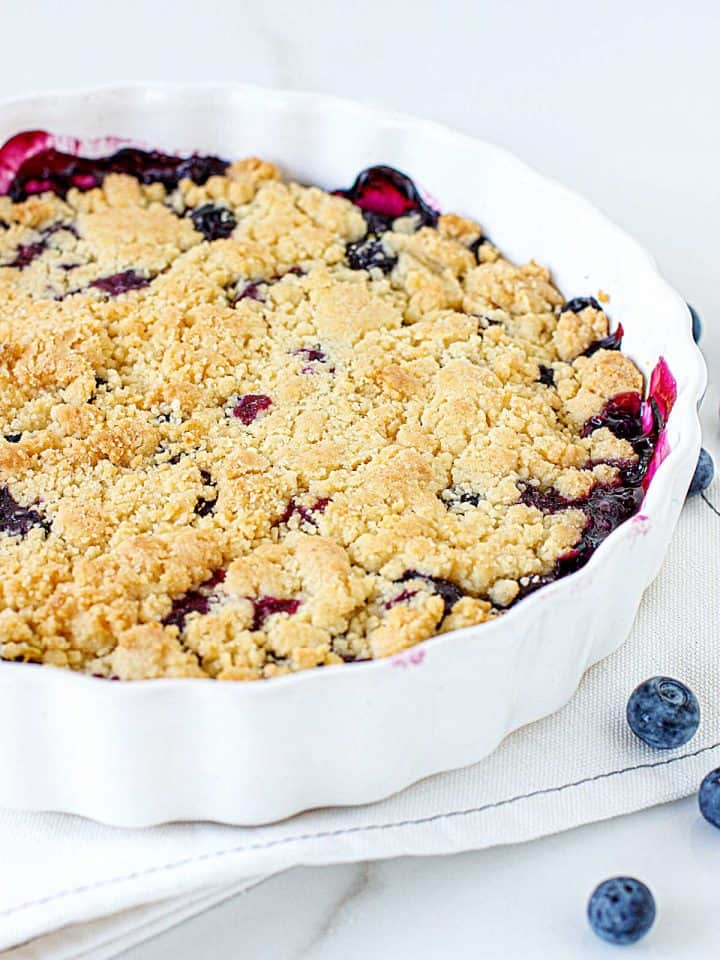 Partial view of round white dish with bluberry crumb cake, white surface, loose blueberries