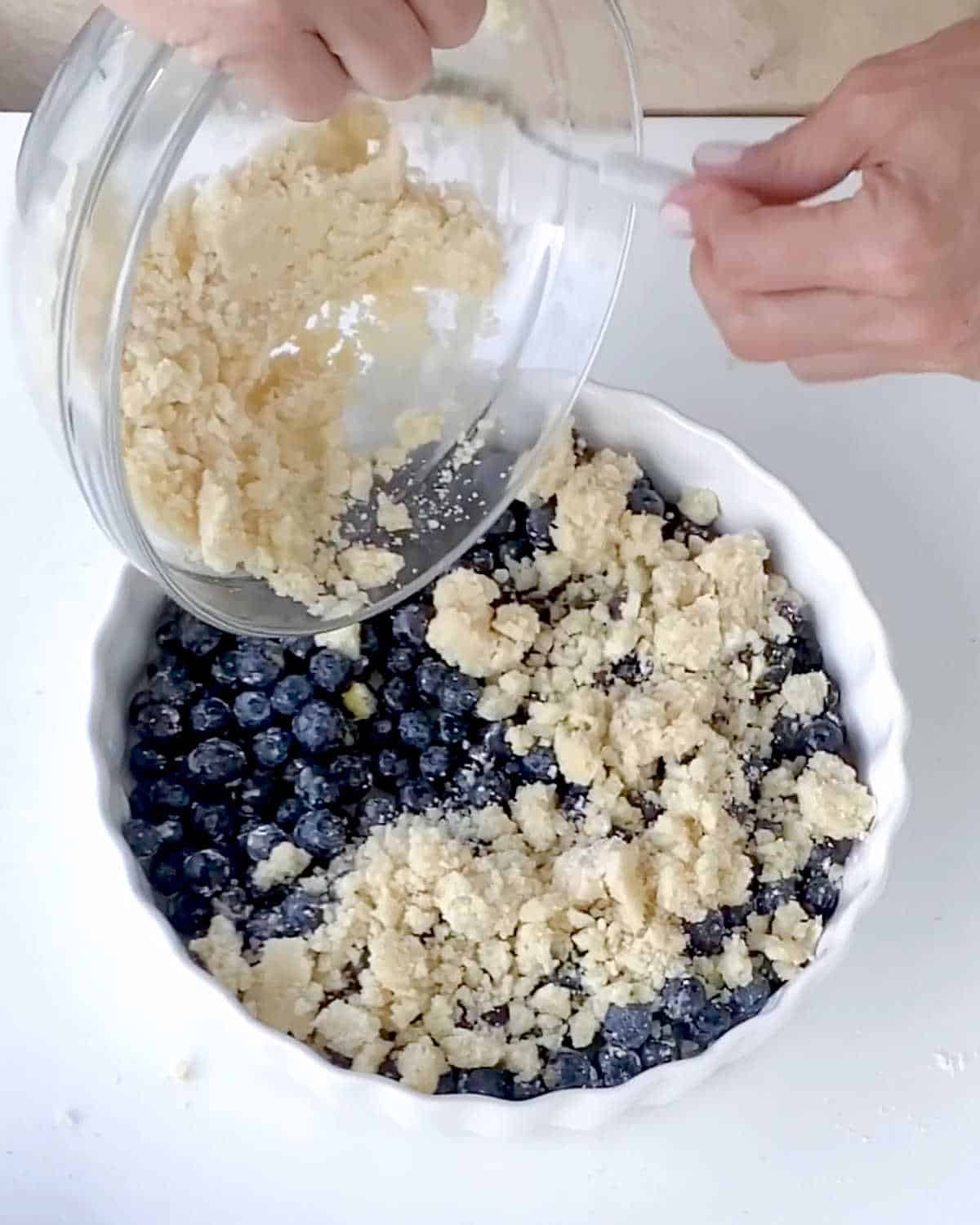 Adding crumbly topping to blueberry dump cake filling in a round white dish on a white table.