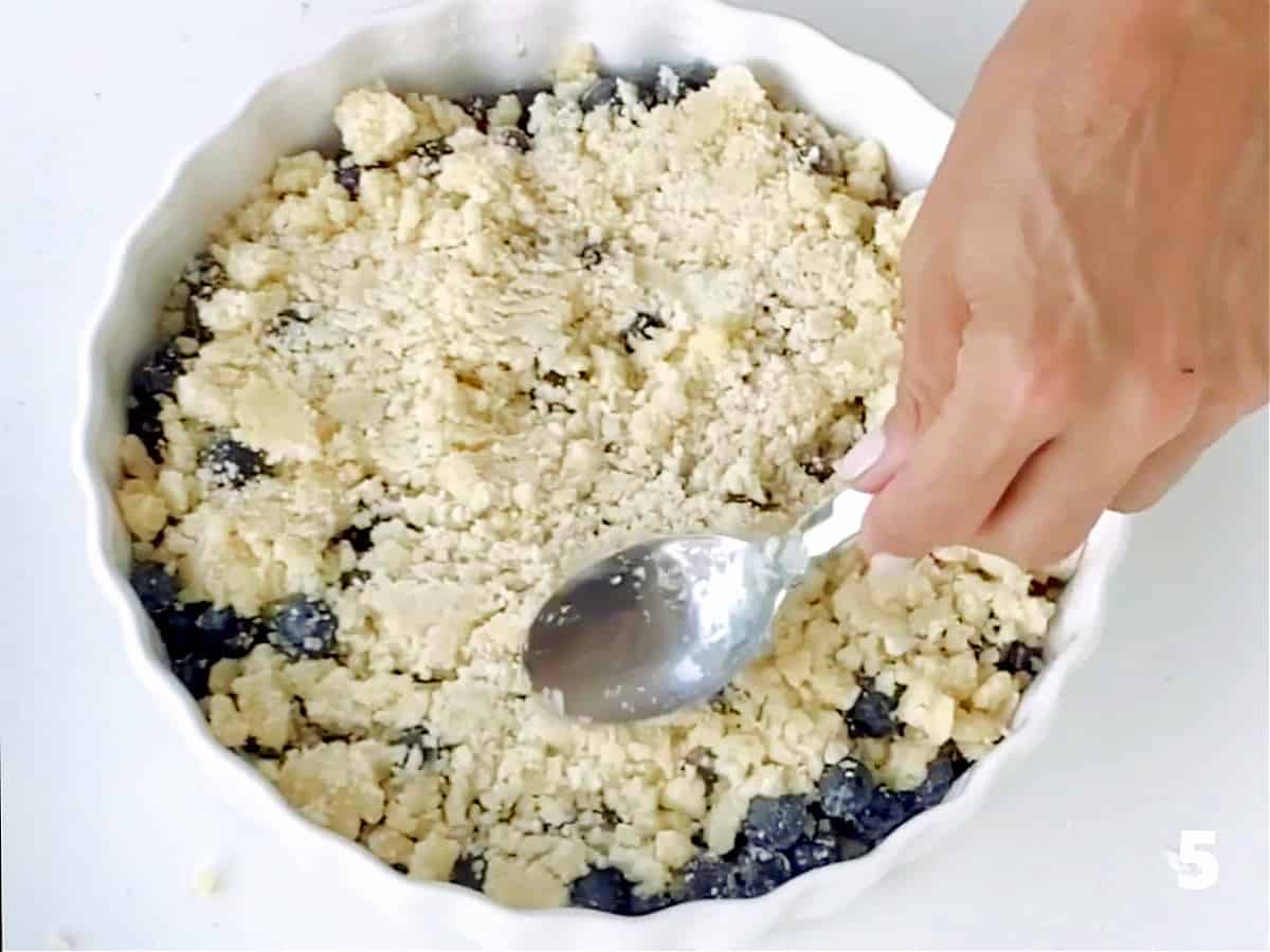 Pressing crumble mixture with spoon on top of blueberries in white ceramic dish on white surface