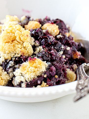 Serving of blueberry dump cake in a white bowl with a silver spoon. White surface.