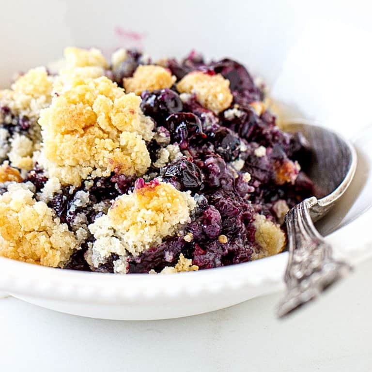 Serving of blueberry dump cake in a white bowl with a silver spoon. White surface.
