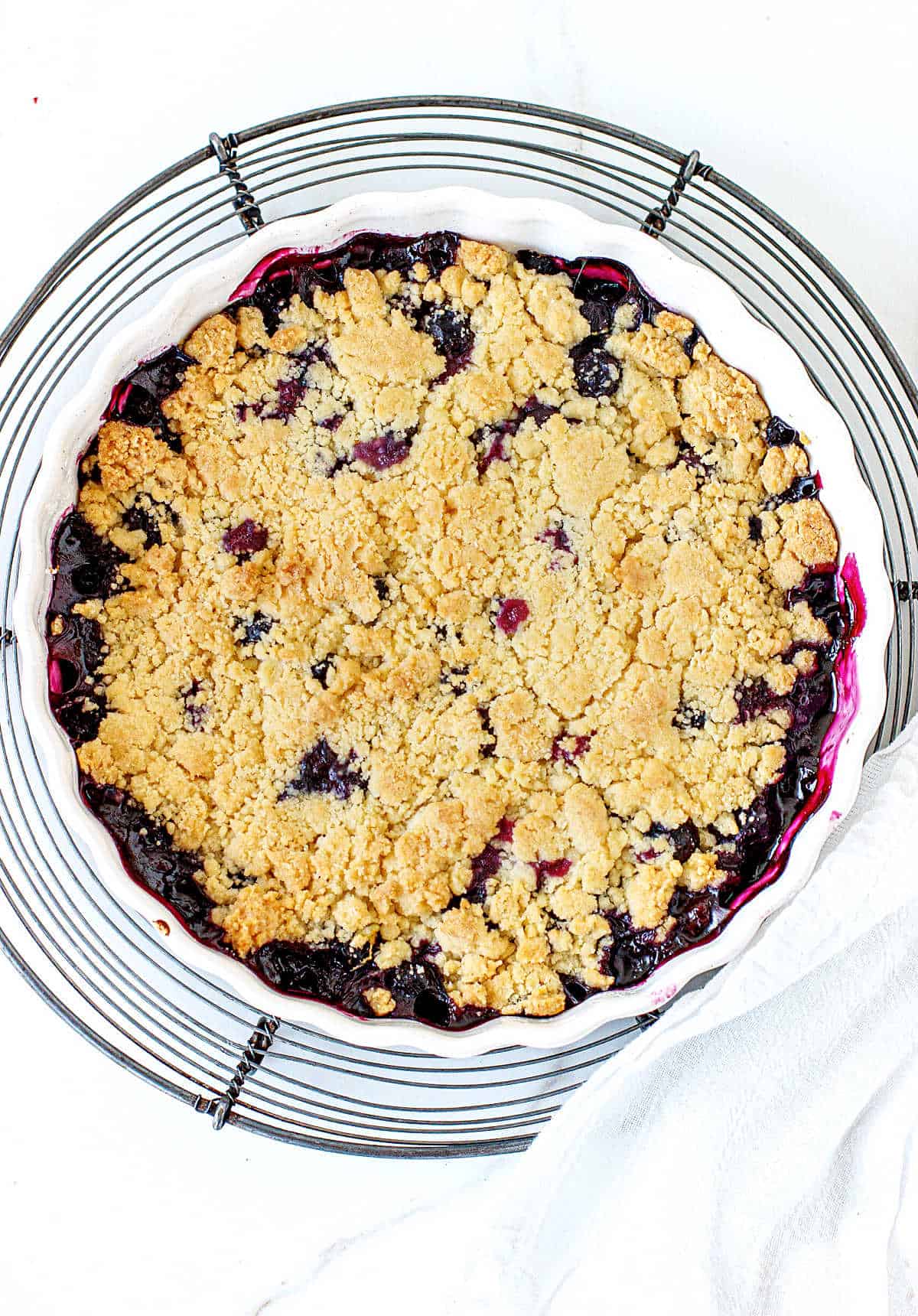 Top view of round white dish with blueberry dessert with crumbly topping on wire rack, white surface and towel.