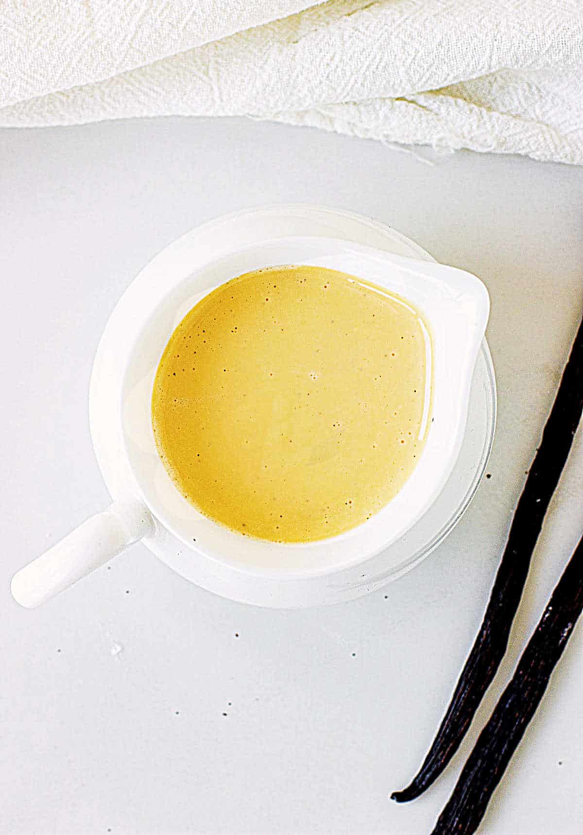 Top view of white jar with custard sauce on a white surface, two vanilla beans.