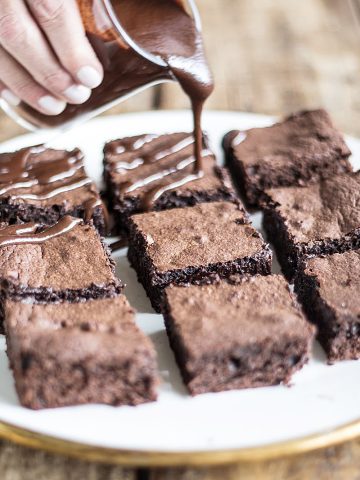Hand pouring chocolate sauce on brownie squares in white plate on wooden table