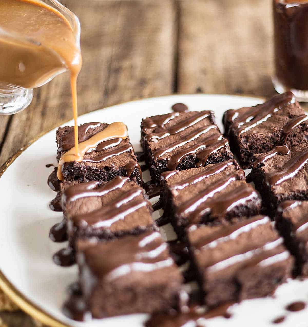Pouring dulce de leche sauce from jar onto brownie squares with chocolate sauce on white plate.