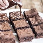 Close up of white plate with fudgy brownies and hand pouring chocolate sauce on top.