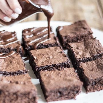 Close up of white plate with fudgy brownies and hand pouring chocolate sauce on top.