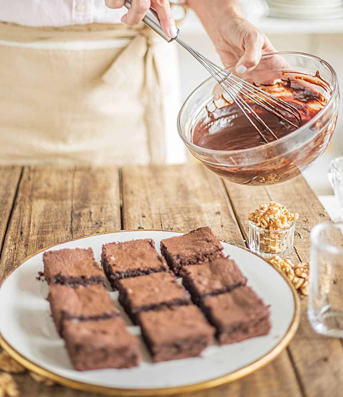 Whisking chocolate sauce in a glass bowl; a plate of brownies below on a wooden table.