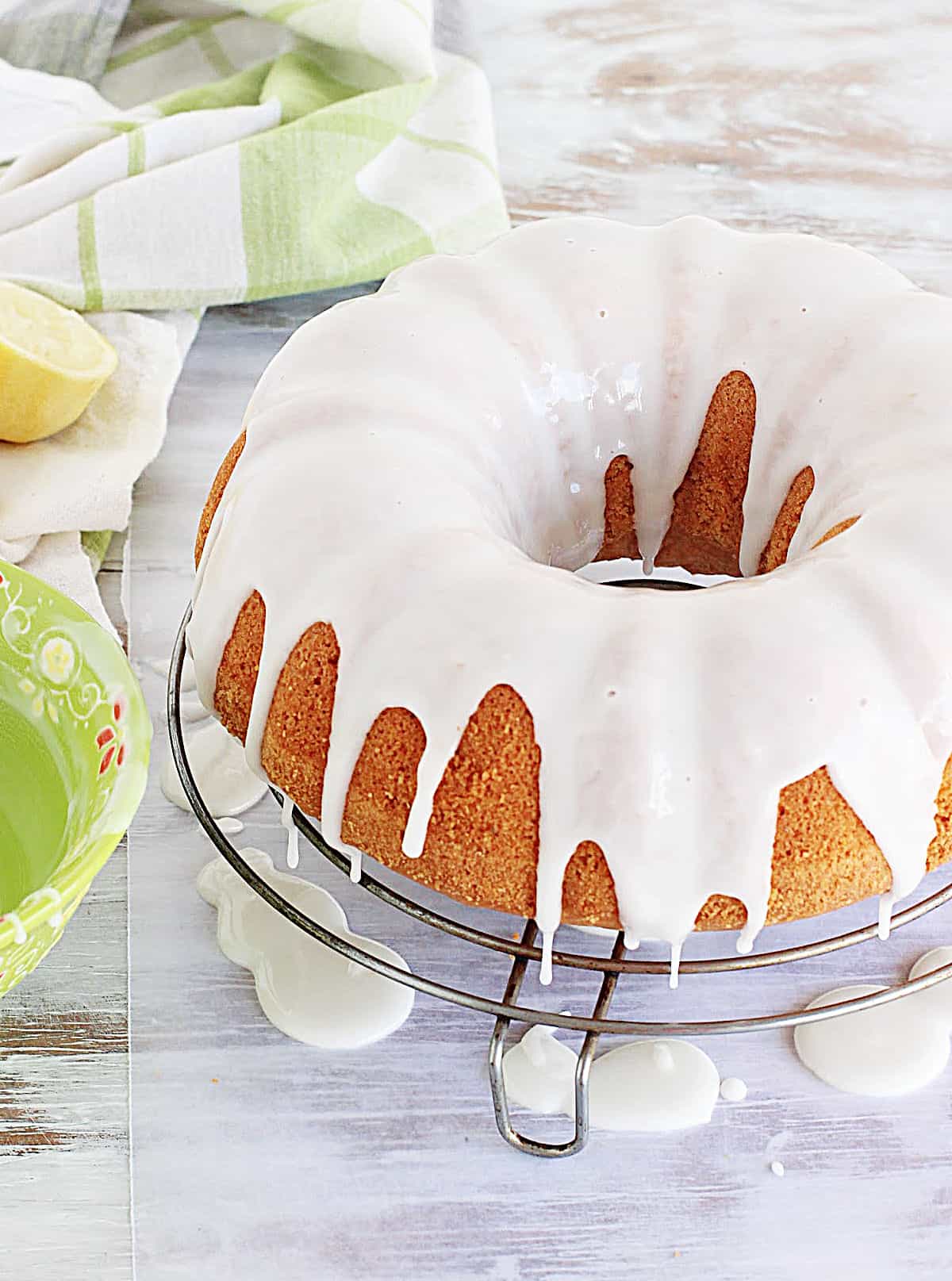 Glazed bundt cake on wire rack on white table, green white cloth and bowl