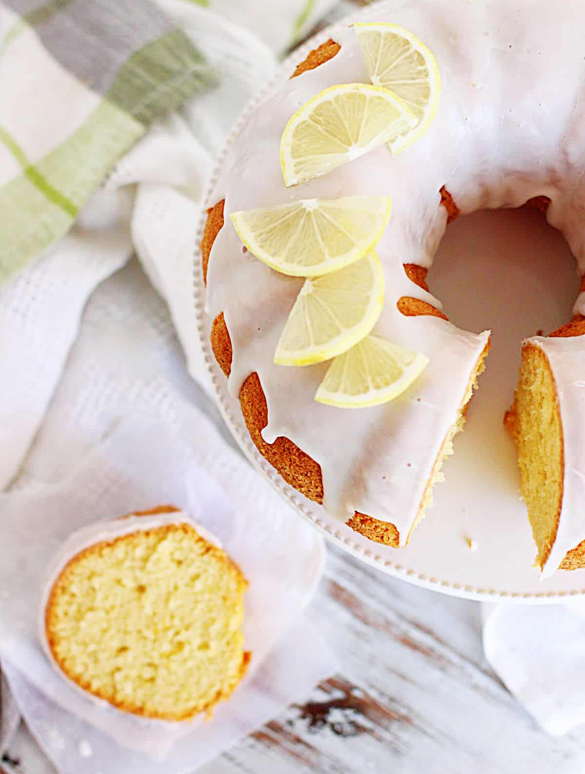 Lemon topped and glazed bundt cake on cake stand, white table, slice of cake and green cloth