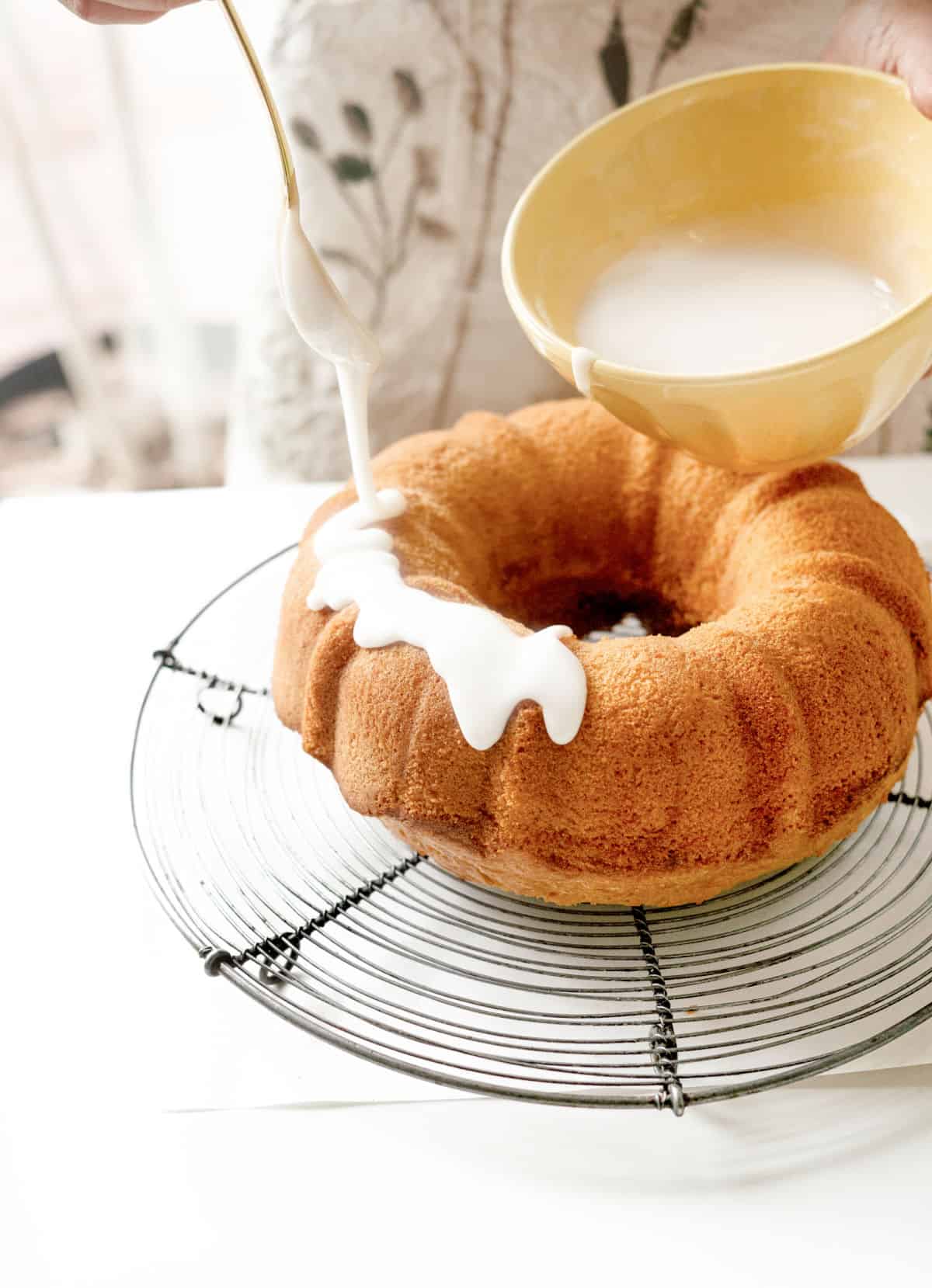 Drizzling a bundt cake with powdered sugar glaze on a wire rack. A yellow bowl.