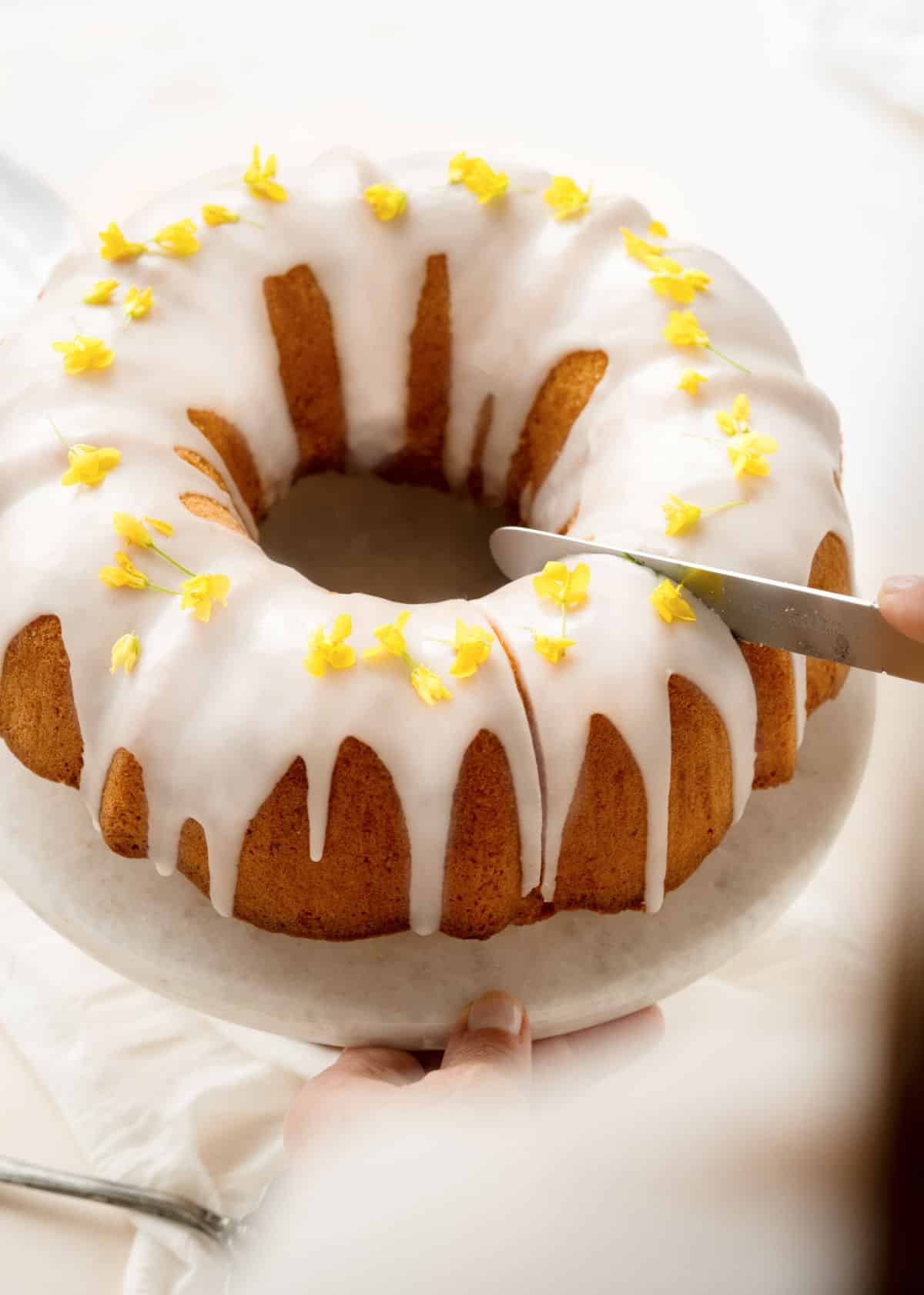 Whole glazed lemon bundt cake being cut with a silver knife on a white background. 