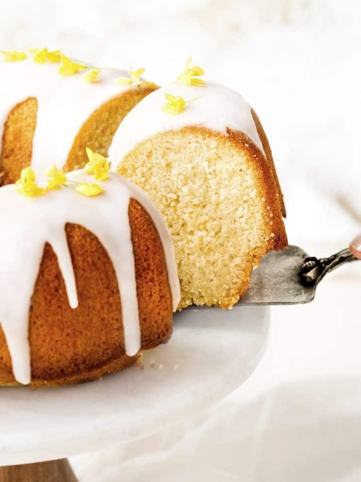 Whole lemon bundt cake on a stand. Hand taking out a slice with a cake server.