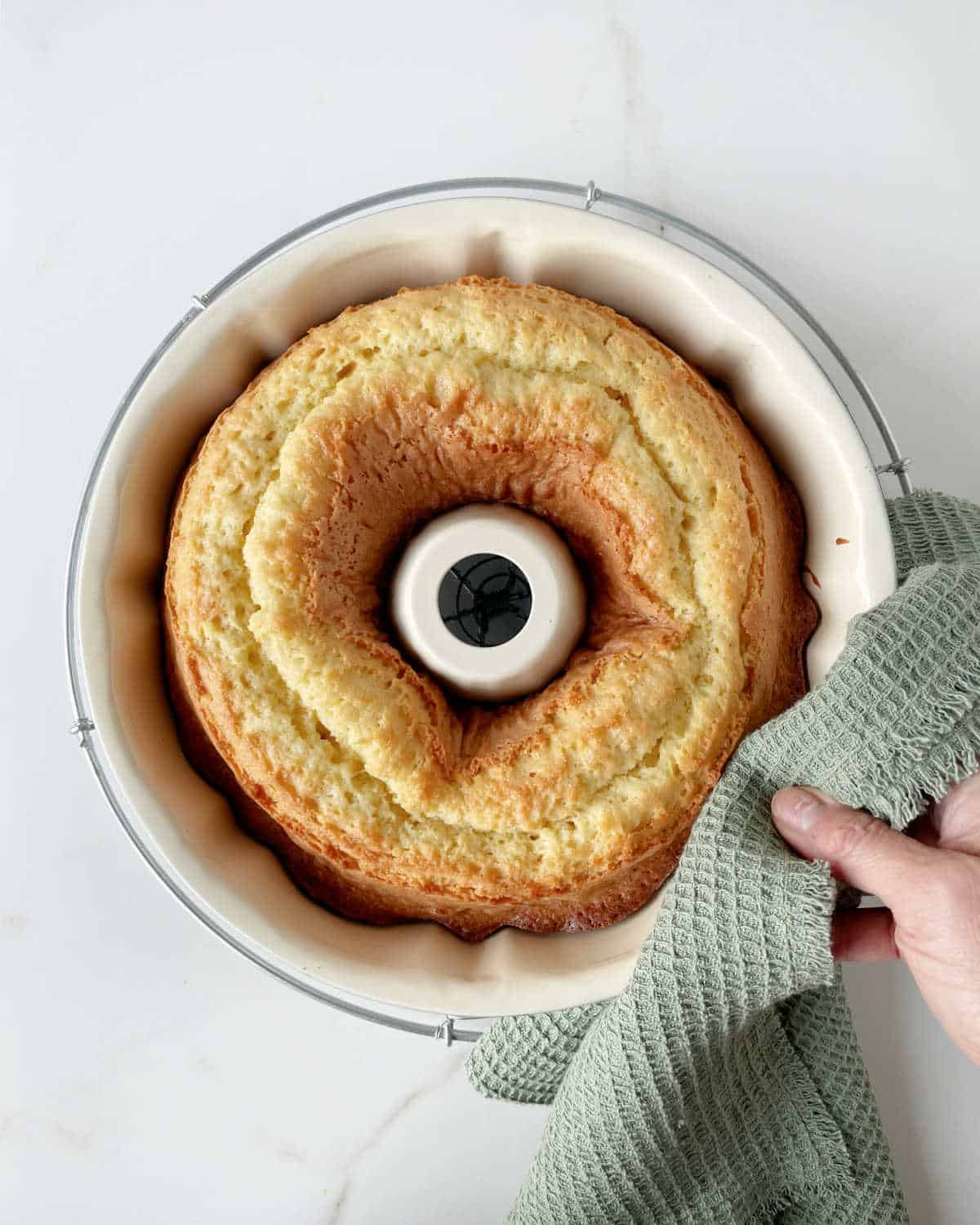 Hand holding baked cake in a bundt pan with a green towel. White surface. 