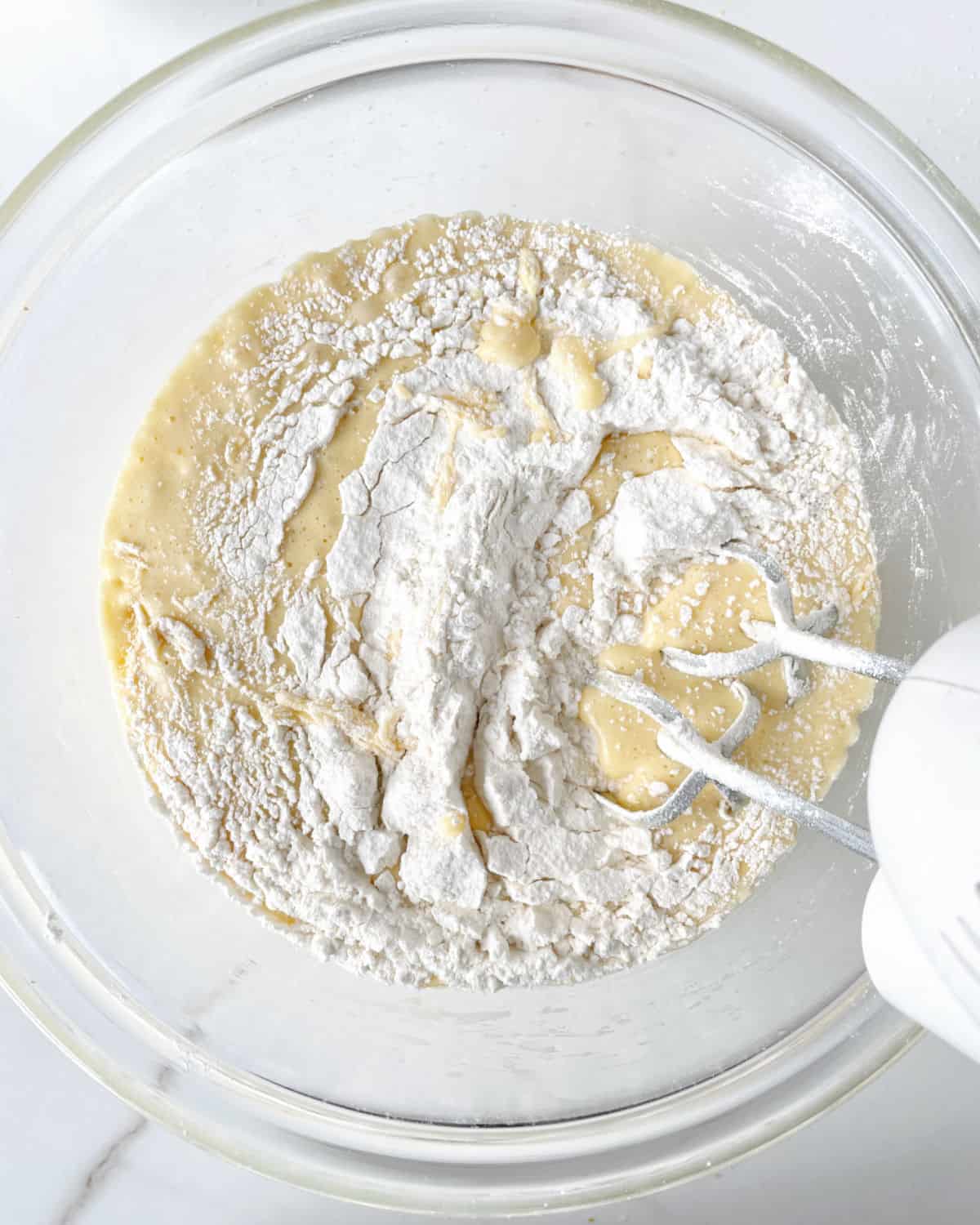 Adding flour to a lemon cake batter in a glass bowl on a white marble surface. 