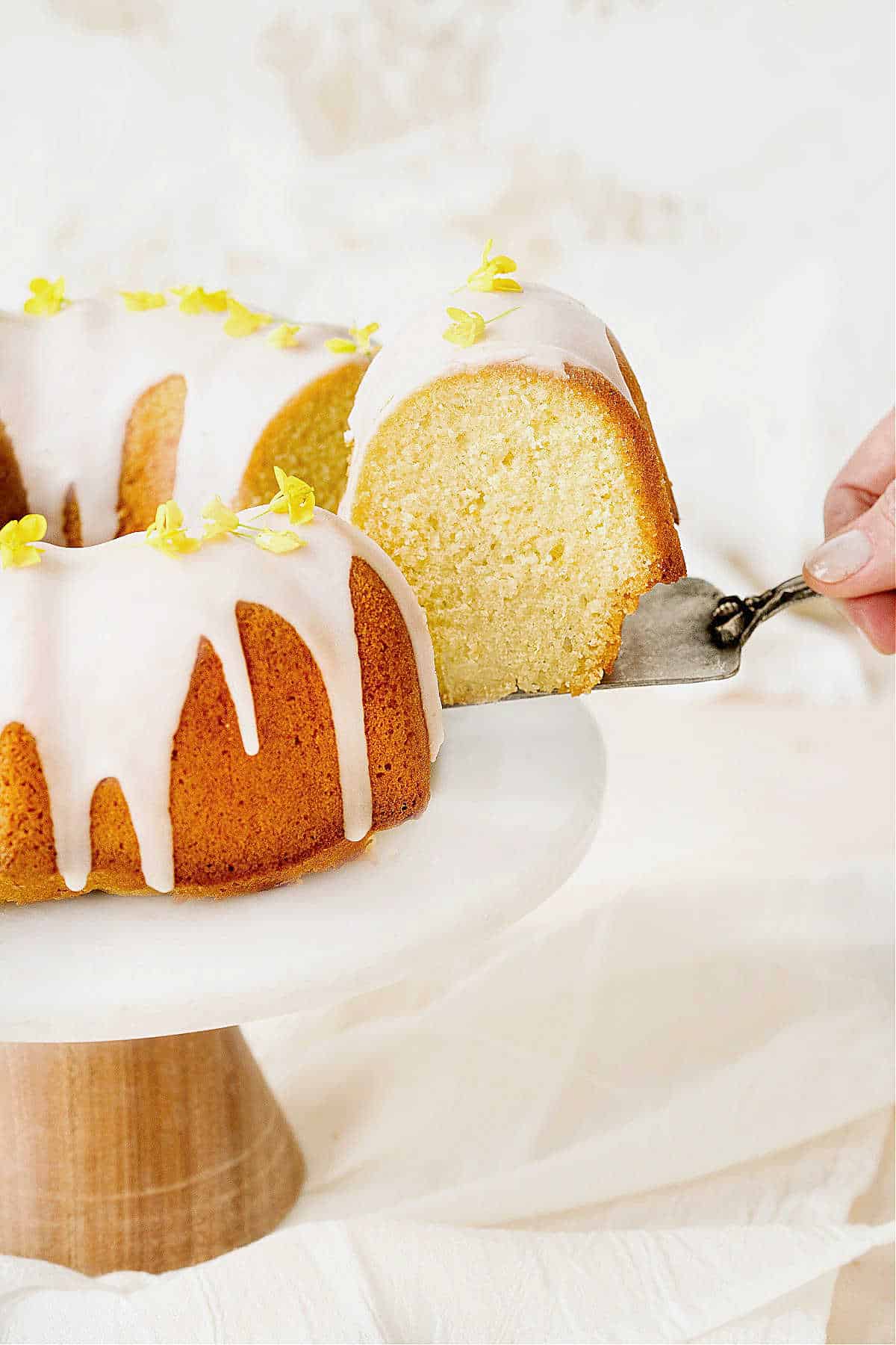 Partial view of glazed lemon bundt cake on a cake stand. A slice being pulled away. White background.