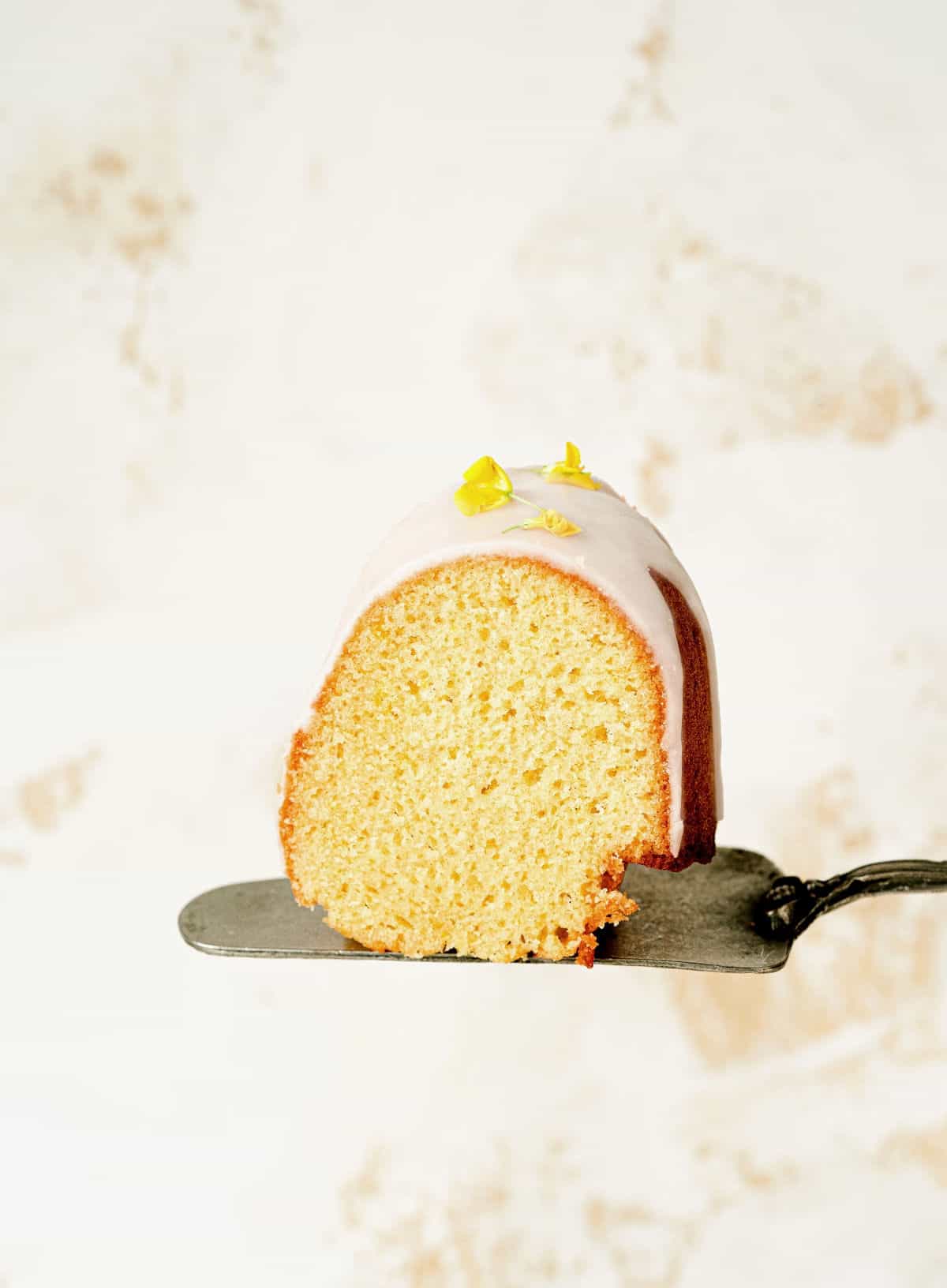 Single slice of glazed lemon bundt cake on a silver cake server. White and beige background.