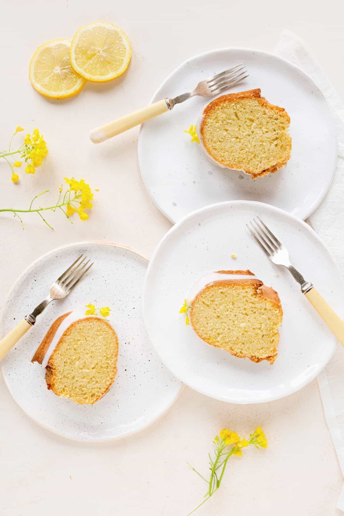 Three white plates with slices of lemon cake, forks, peach colored surface, yellow flowers and lemon slices. Top view.