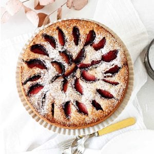 View from above of whole plum cake on a white kitchen towel. Yellow forks beside it.