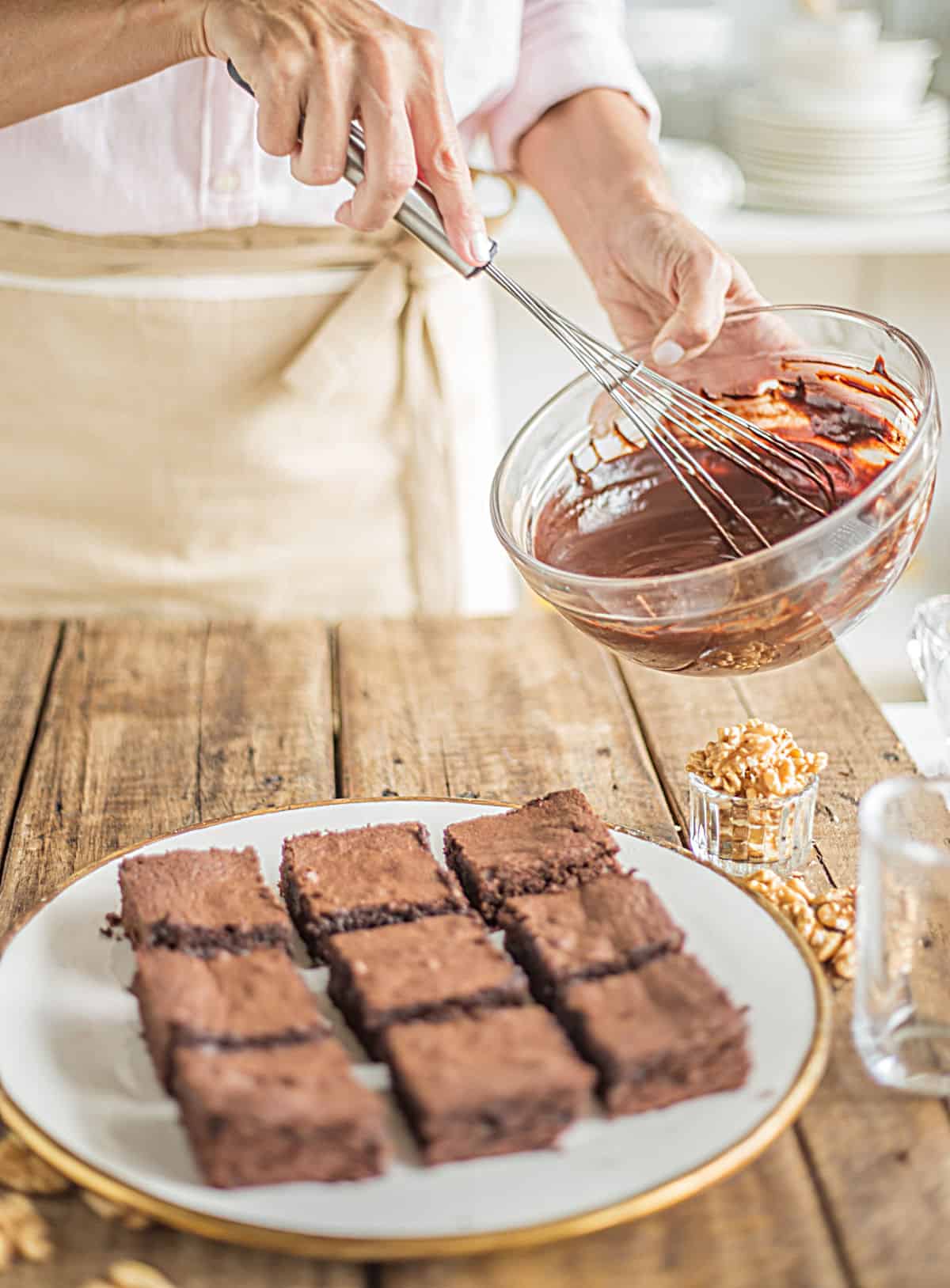 Person with pink and beige apron beating chocolate sauce with plate of brownies below on wooden table