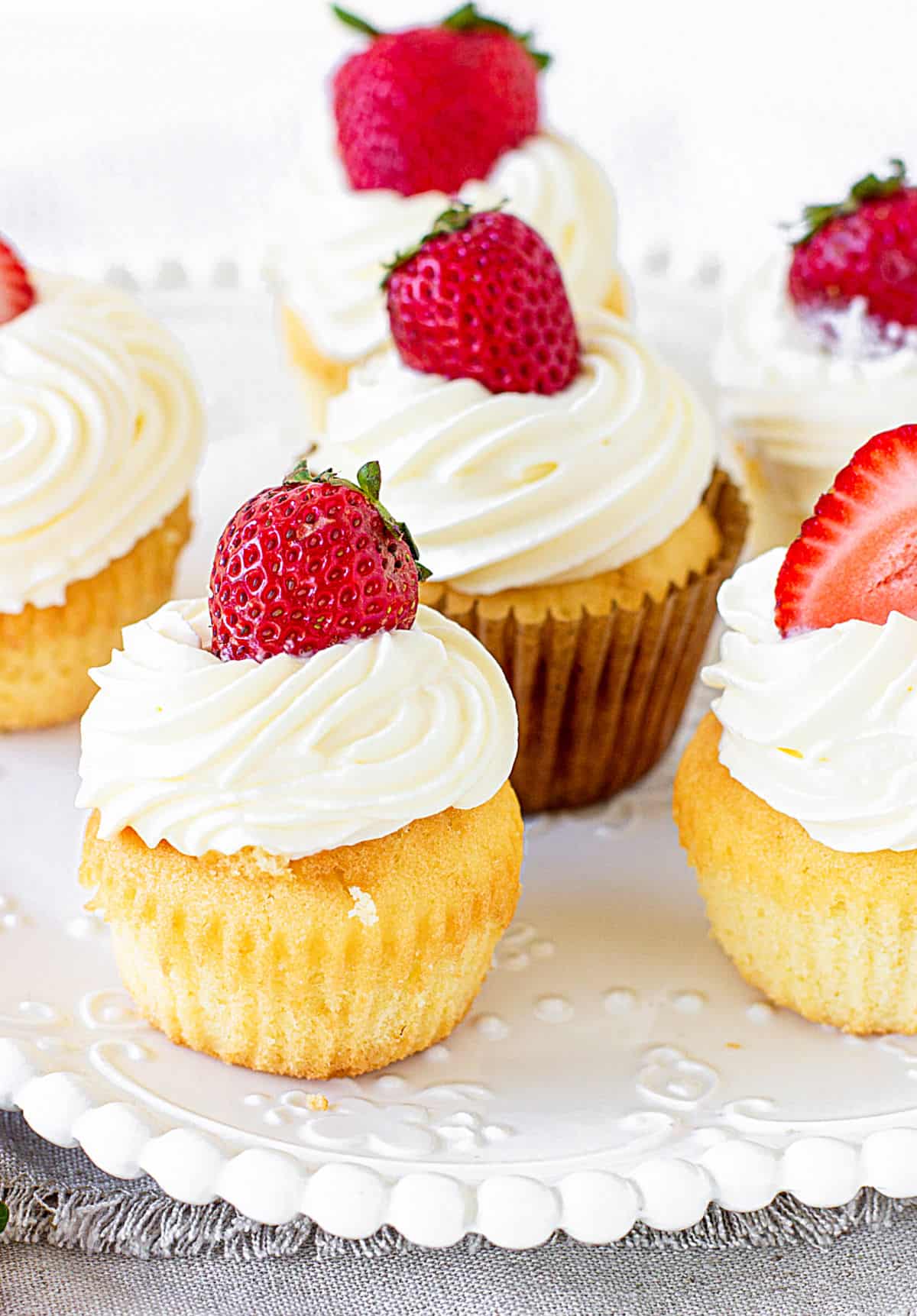 White plate with several cream and strawberry topped vanilla cupcakes