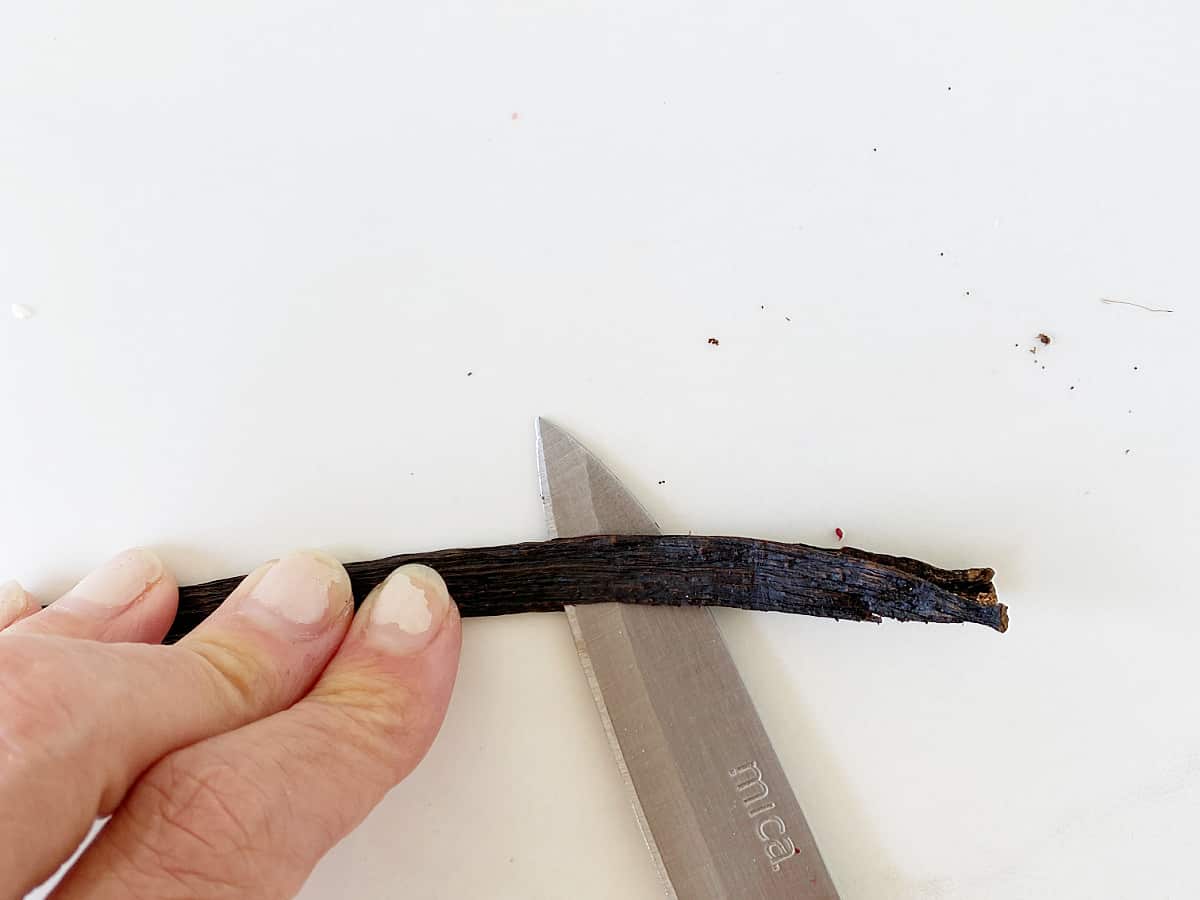 Opening vanilla bean with knife, hand holding it down, white surface