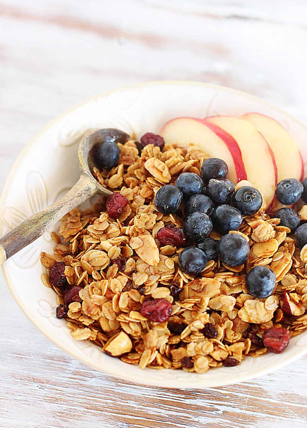 White bowl filled with granola, apples and blueberries, with wooden spoon, on white table