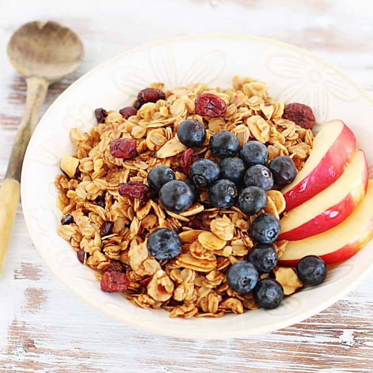 Whitish table with white bowl with granola and fruit, a wooden spoon beside it.