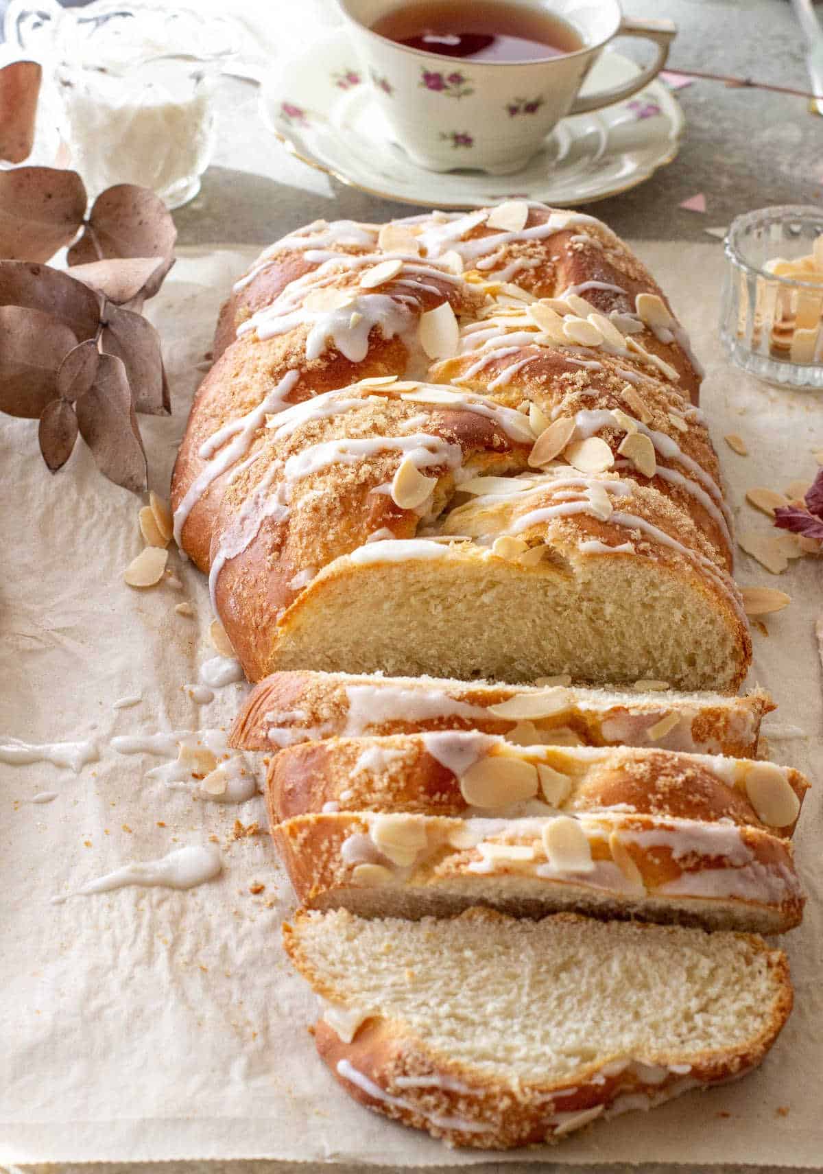 A loaf of glazed braided Easter Bread with sliced almonds. Beige surface. A cup of tea in the background.