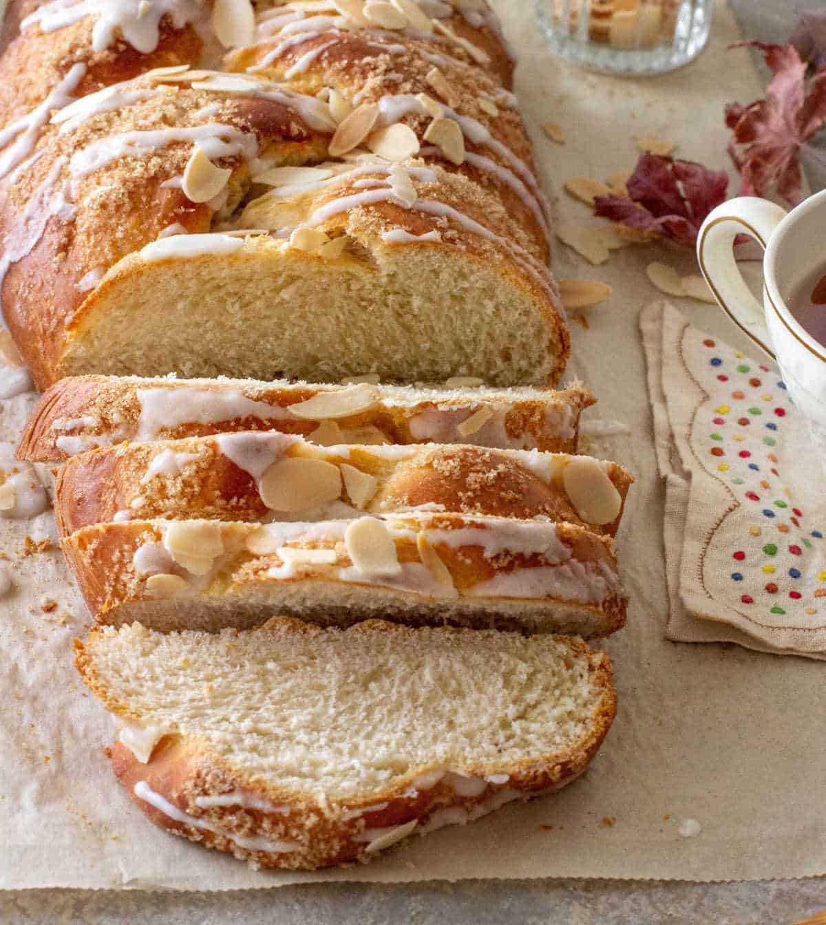 Sliced glazed braided almond Easter bread on a beige paper. Napkin, cup, and dry leaves around. 