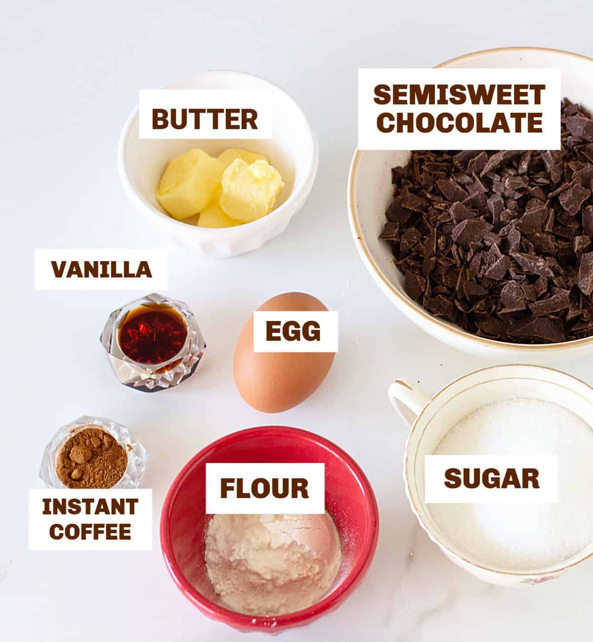 Different bowls with ingredients for brownie cookies including egg, chopped chocolate, flavorings, butter on white surface