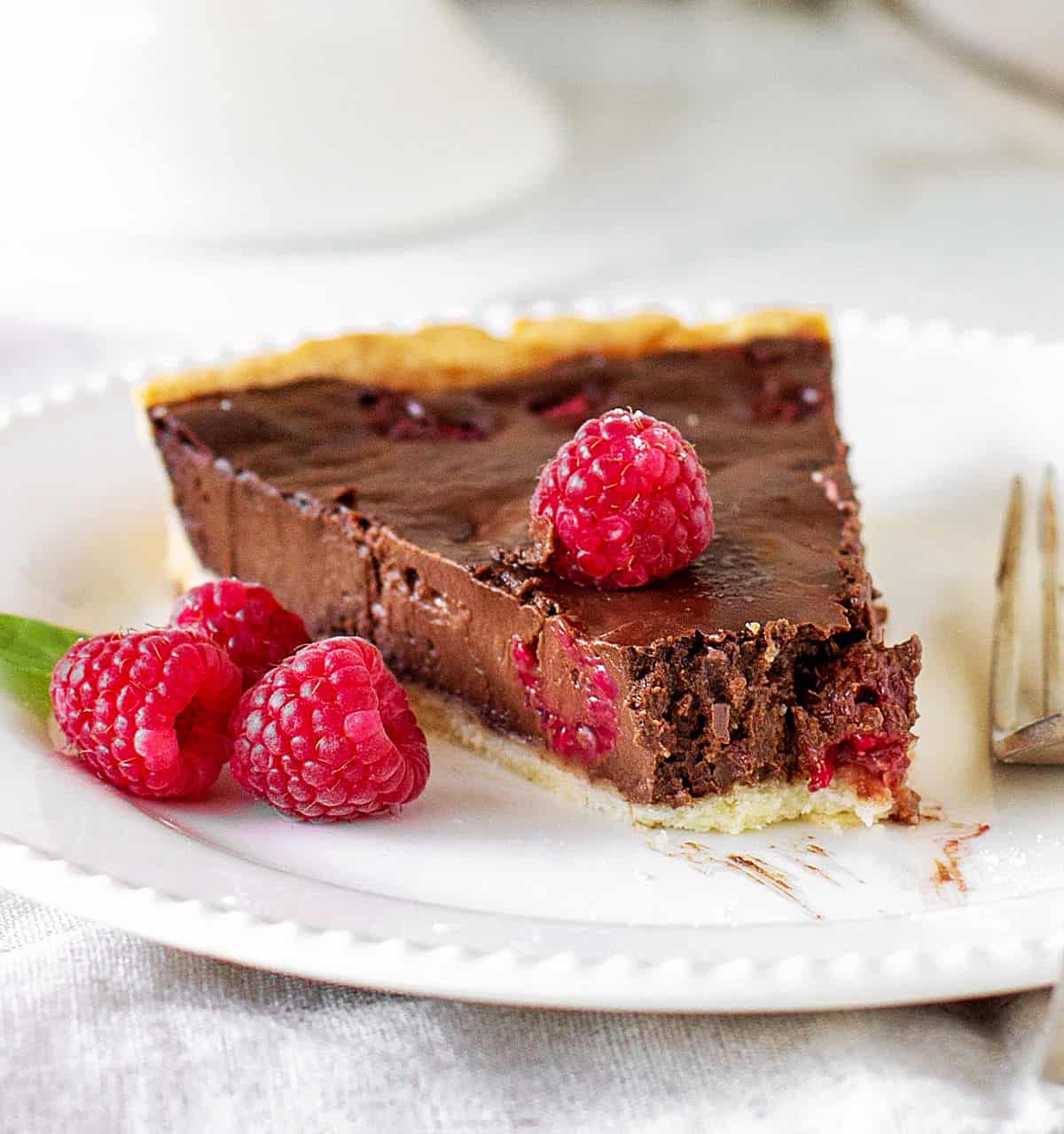 Slice of chocolate raspberry tart with missing bite, on white plate, silver fork.