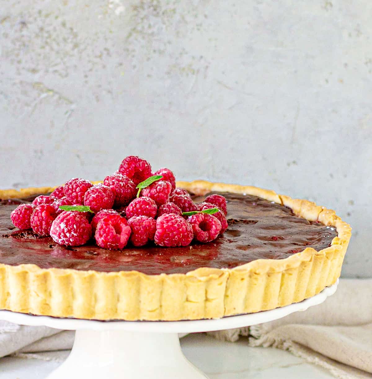 Partial image of chocolate tart on white cake stand, whole raspberries on top