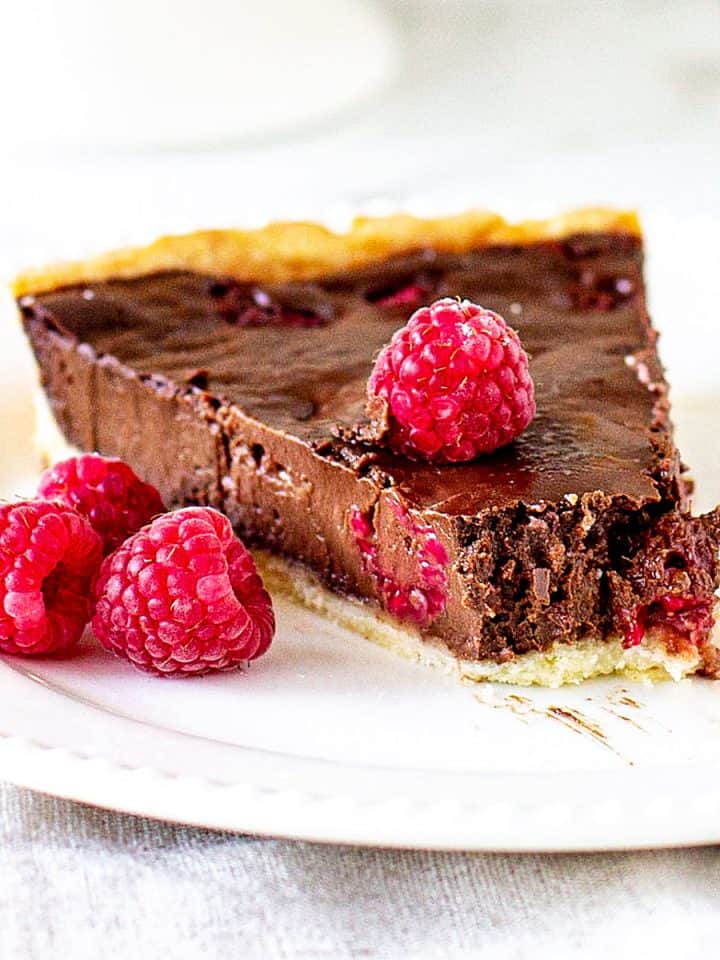 Single slice of raspberry chocolate tart on white plate, whole raspberries, white background.
