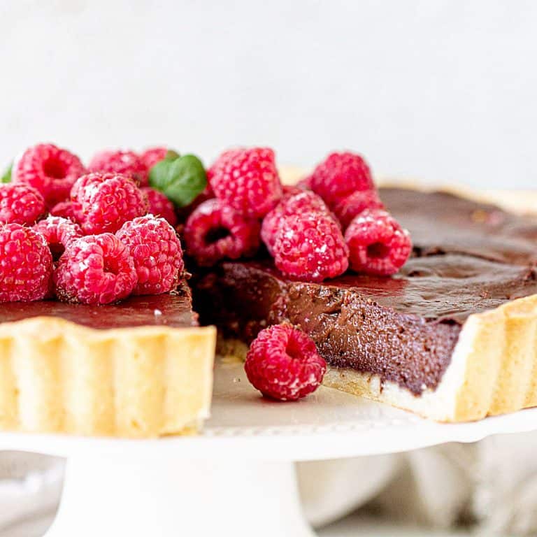 Close up of chocolate tart on a white cake stand with a missing slice. Fresh raspberries on top. White background.