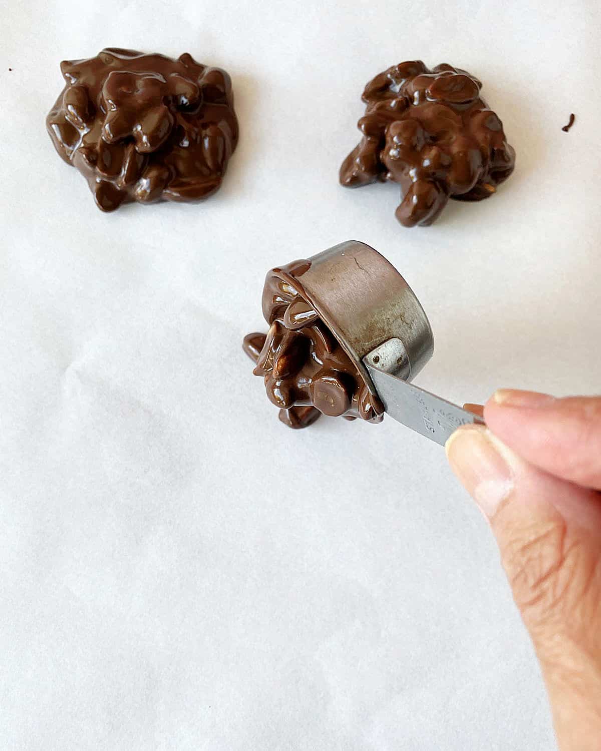 Mounding chocolate peanut clusters with small measuring cup on parchment paper.