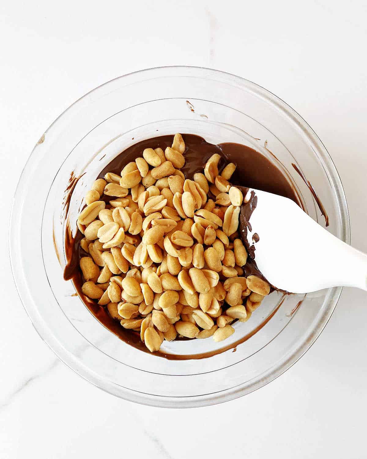 Peanuts added to melted chocolate in a glass bowl with a white spatula. White marble suface.