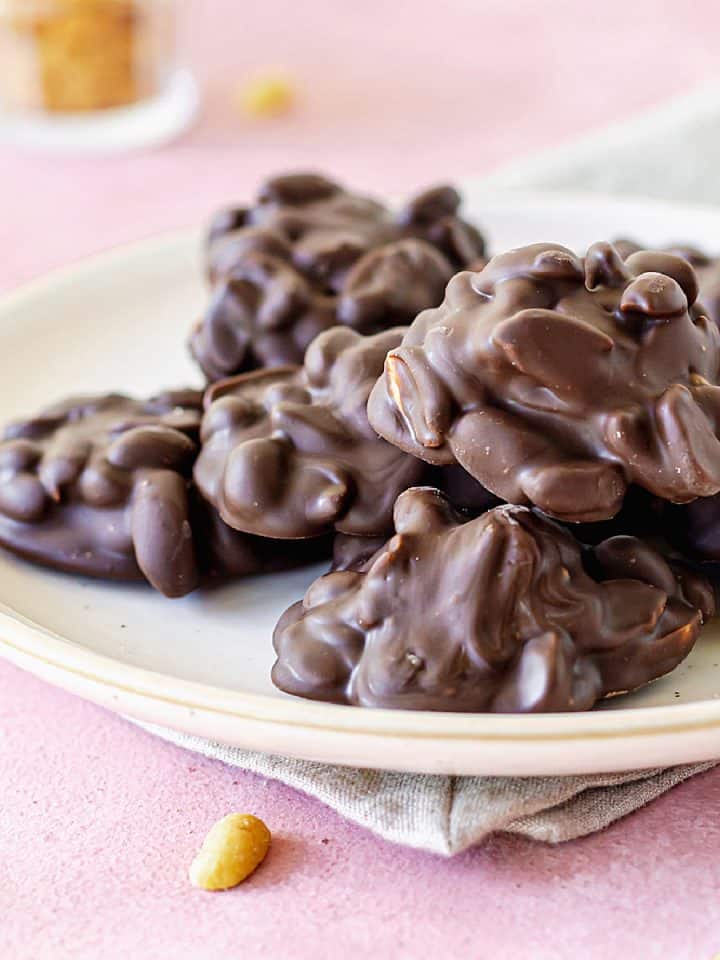 Pile of chocolate peanut clusters on a white plate set on a pink surface with loose peanuts around.