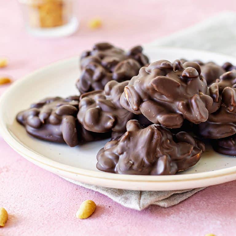 Pile of chocolate peanut clusters on a white plate set on a pink surface with loose peanuts around.