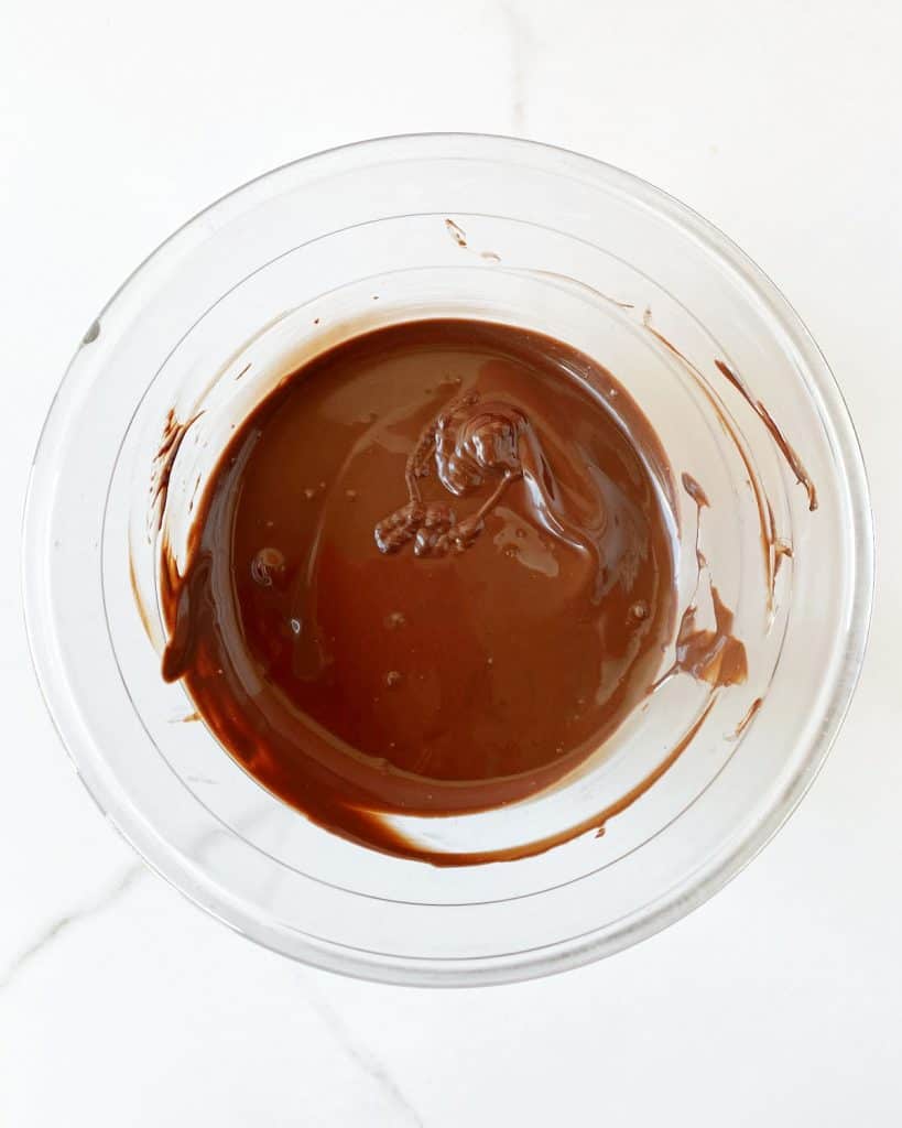 Melted dark chocolate in a glass bowl on a white marble surface.