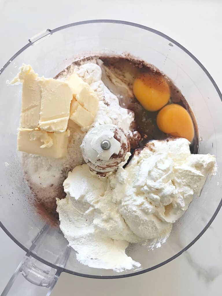 Bowl of food processor with sour cream, butter, eggs, cocoa powder and flour mixture. Top view.