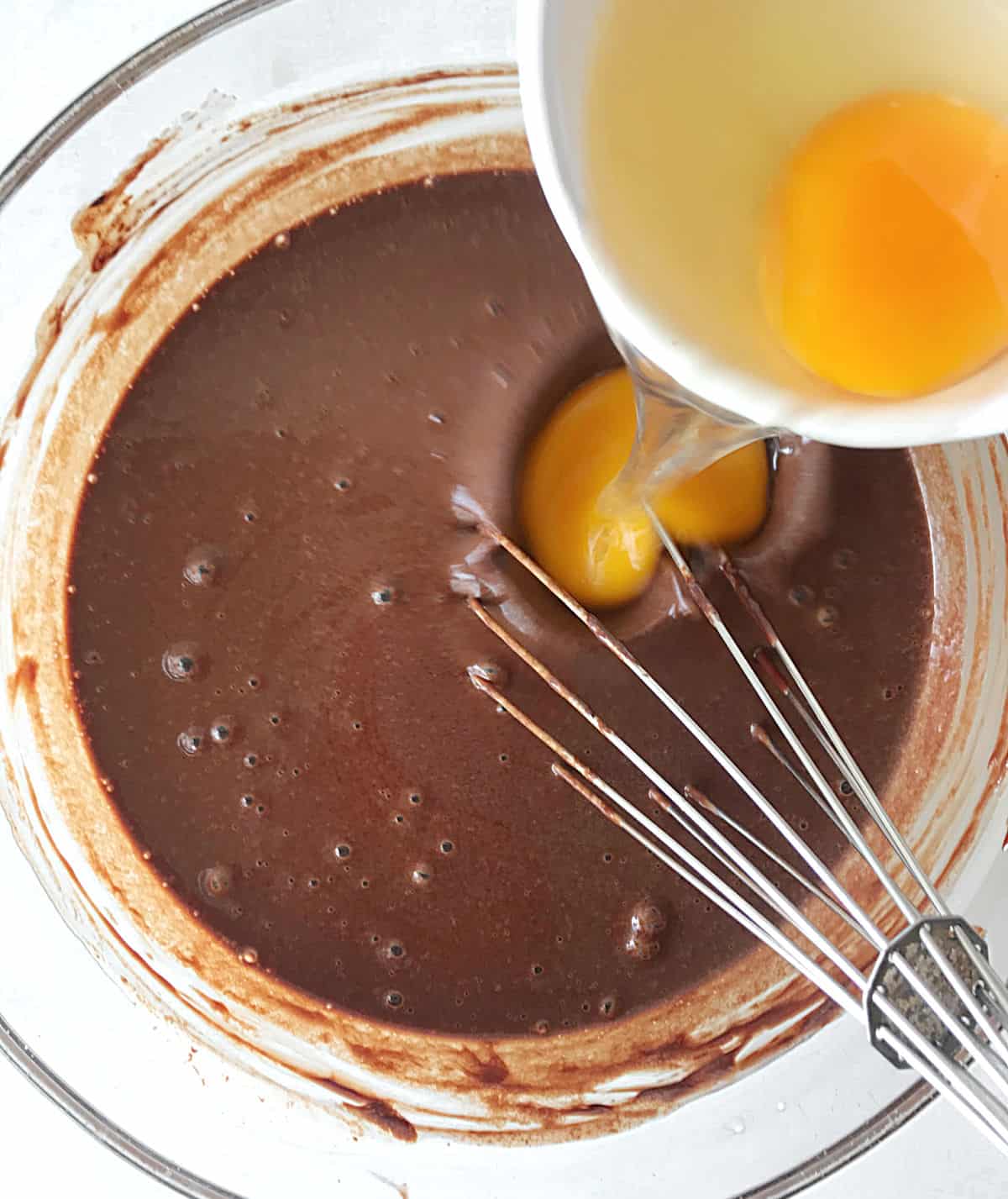 Adding eggs to chocolate mixture in a glass bowl with a whisk inside. White surface, top view.