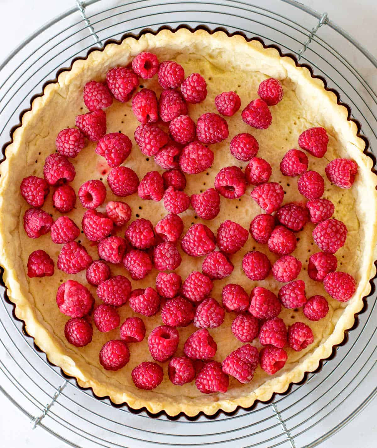 Fresh raspberries in a tart shell on a wire rack on a white marble surface.