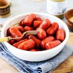 Silver fork pinching mini sausage among several in white bowl, grey linen and wooden board underneath.
