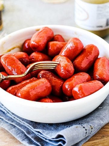 Silver fork pinching mini sausage among several in white bowl, grey linen and wooden board underneath.