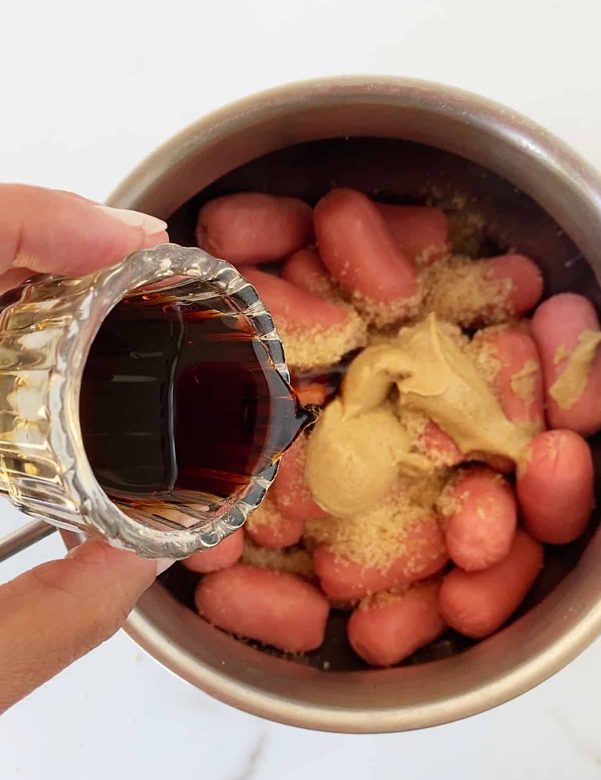 Adding soy sauce to metal saucepan with cocktail sausages on white marble surface