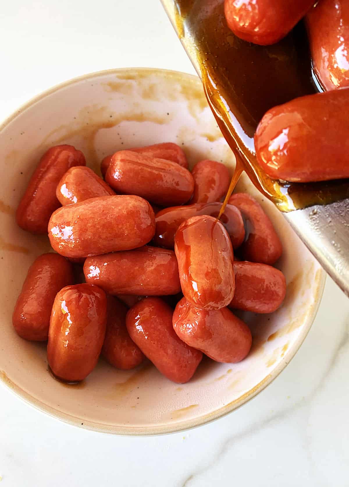 Transferring little sausages with syrup to white bowl on white surface